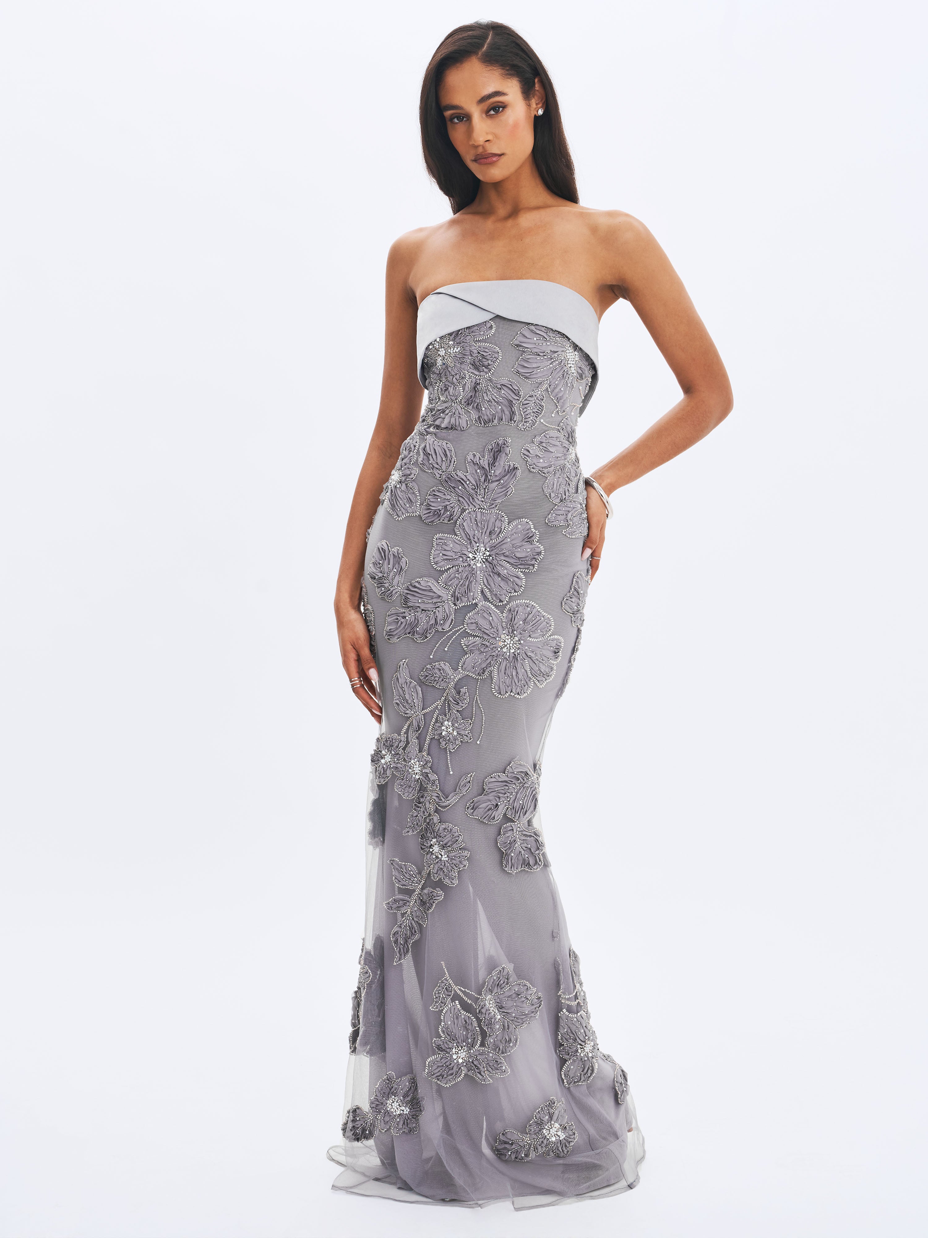 Woman wearing a strapless, silver evening gown with floral embroidery on a white background